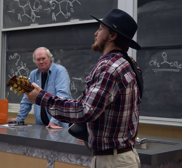 On the right, a male student with a beard in a cowboy hat and plaid shirt plays the guitar and sings. On the left in the background, an older man stands watching and smiling. He is wearing a demin shirt unbuttoned over a black tshirt.