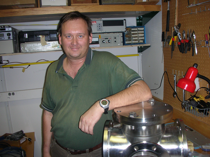 Dr. Sergei Aksyonov stands in the back corner of a lab surrounded by equipment and smiles at the camera