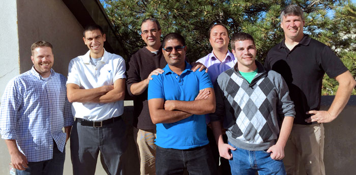 Seven members of the antibiofilm antibiotic research project from Prof. Ryan Loopers group stand together in a group, smiling at the camera. They are outside with a pine tree in the background.