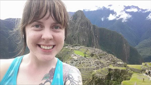 A young woman in a tank top with a shoulder tattoo smiles at the camera, with Machu Picchu far in the background