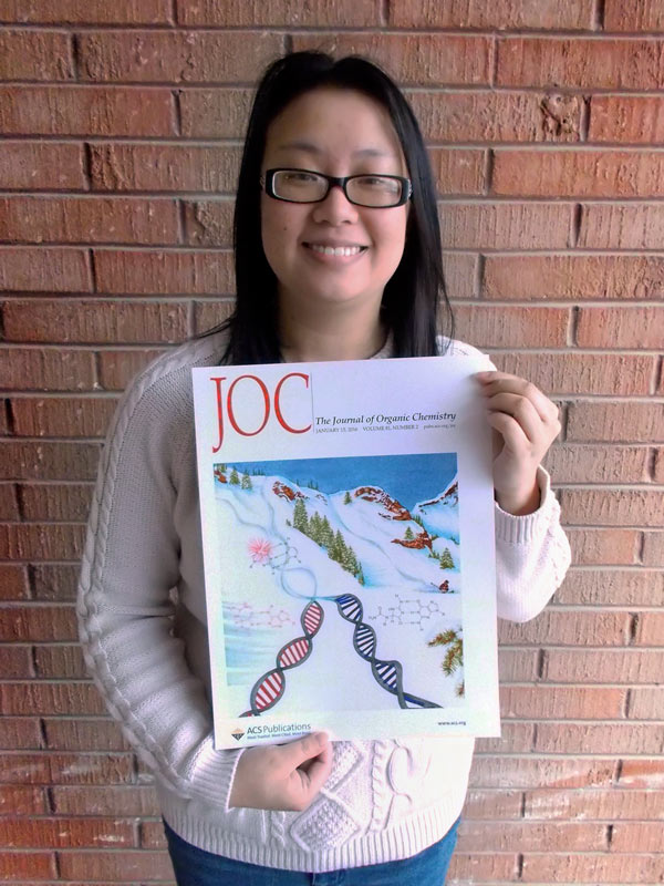 Judy Zhu stands outside in front of a brick wall. She has long dark hair around her shoulders and is wearing a white sweater and jeans. She is holding the cover of the Journal of Organic Chemistry, which shows an illustration of a snowy slope with two ski tracks that diverge into two DNA strands, one red and one blue. Around the DNA strands are structures of the molecules studied in Zhu's paper.